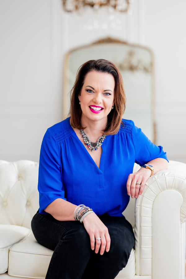 Empowering woman smiling in elegant blue blouse sitting on white sofa, Marie Monville, inspirational speaker and author promoting faith, hope, and resilience.
