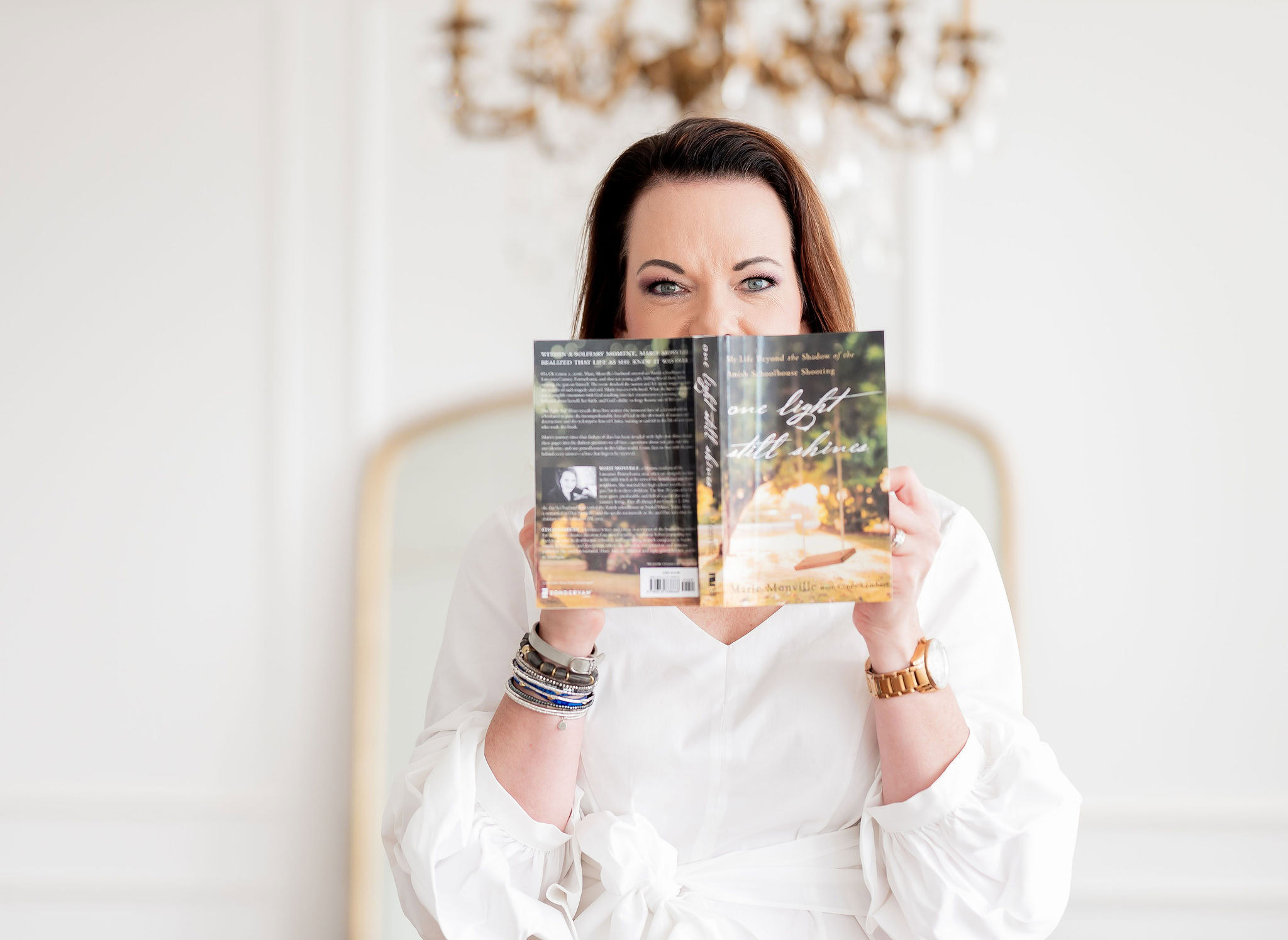 A woman with dark hair holds a book titled "One Light Still Shining" in front of her face, indoors with elegant decor, reflecting inspiration and resilience, emphasizing personal growth and hope.