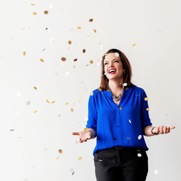 Marie Monville celebrating with gold and white confetti, showcasing joy and positivity, indoor setting, dressed in a vibrant blue blouse, emphasizing inspiration and resilience.