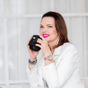 Warm professional woman smiling with coffee mug, wearing white blazer and jewelry, in bright modern office setting.